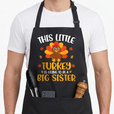 Sister thanksgiving apron - Cartoon turkey with pink bow and six autumn leaves surrounding big sister announcement text — shown on a black cotton canvas apron against a white background