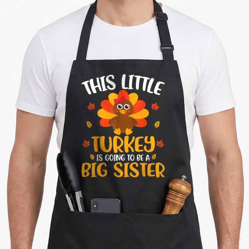 Sister thanksgiving apron - Cartoon turkey with pink bow and six autumn leaves surrounding big sister announcement text — shown on a black cotton canvas apron against a white background