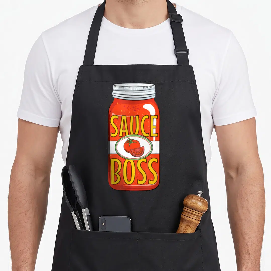 Sauce pasta apron - Cartoon red sauce jar with SAUCE BOSS text and tomato illustration — shown on a black cotton canvas apron against a white background