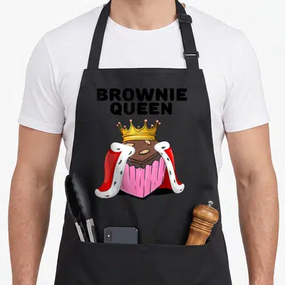 Brownie queen apron - Cartoon brownie wearing gold crown and red white royal cape under stacked BROWNIE QUEEN text — shown on a black cotton canvas apron against a white background