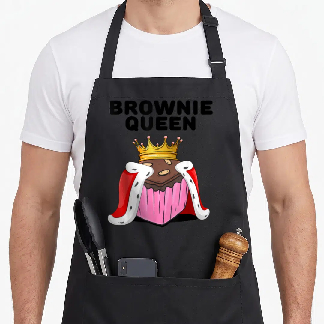 Brownie queen apron - Cartoon brownie wearing gold crown and red white royal cape under stacked BROWNIE QUEEN text — shown on a black cotton canvas apron against a white background