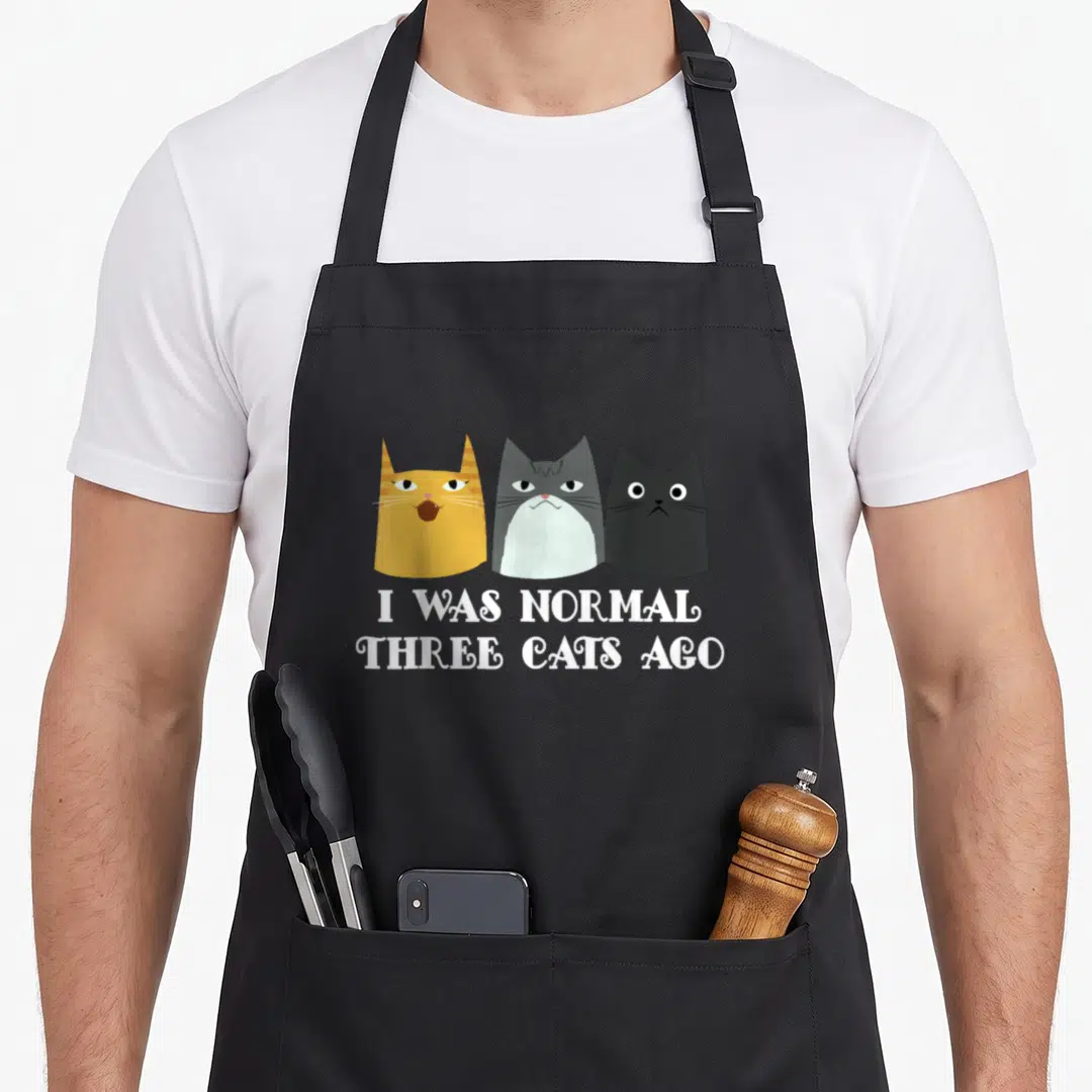 Ago cat apron - Three cartoon cats in orange grey and black above white text I Was Normal Three Cats Ago — shown on a black cotton canvas apron against a white background