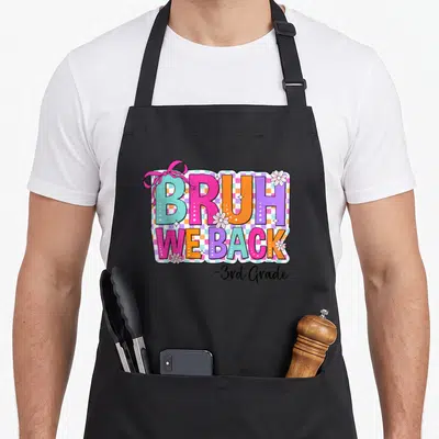 Grade bow apron - Colorful BRUH WE BACK text with pink bow and white flowers on checkered background — shown on a black cotton canvas apron against a white background