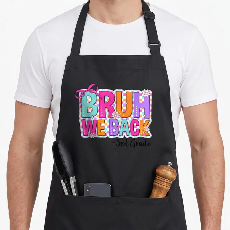 Grade bow apron - Colorful BRUH WE BACK text with pink bow and white flowers on checkered background — shown on a black cotton canvas apron against a white background