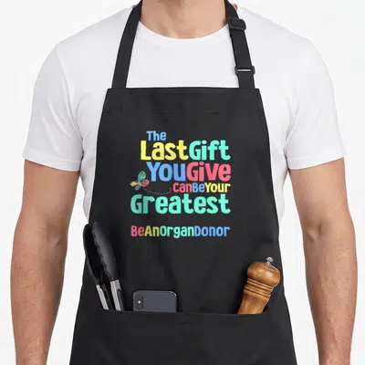 Donor donation organ apron - Stacked colorful text reading The Last Gift You Give Can Be Your Greatest — shown on a black cotton canvas apron against a white background