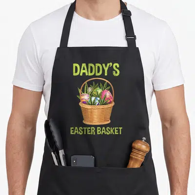 Daddy basket apron - Cartoon basket with colorful eggs and green leaves labeled Daddy's Easter Basket — shown on a black cotton canvas apron against a white background