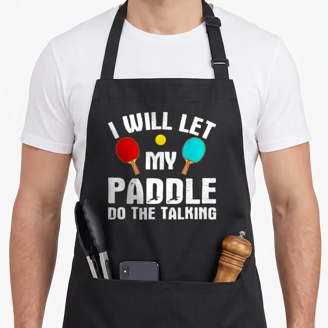 Tennis paddle table apron - White stacked text reading I will let my paddle do the talking above red and blue paddles — shown on a black cotton canvas apron against a white background