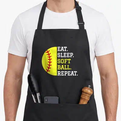 Eat softball player apron - Half-yellow softball with red stitching beside stacked eat sleep softball repeat text — shown on a black cotton canvas apron against a white background