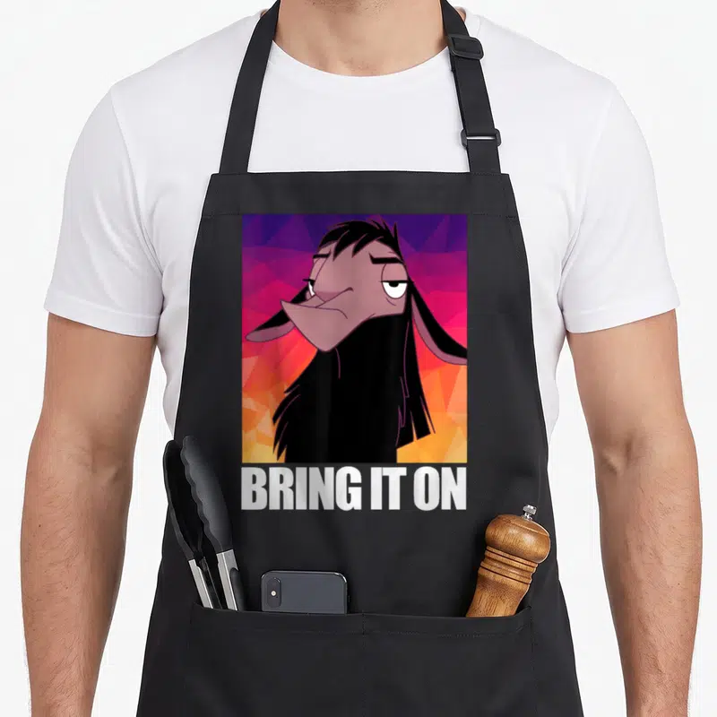 Groove llama apron - Cartoon goat portrait with BRING IT ON text on geometric purple and orange background — shown on a black cotton canvas apron against a white background