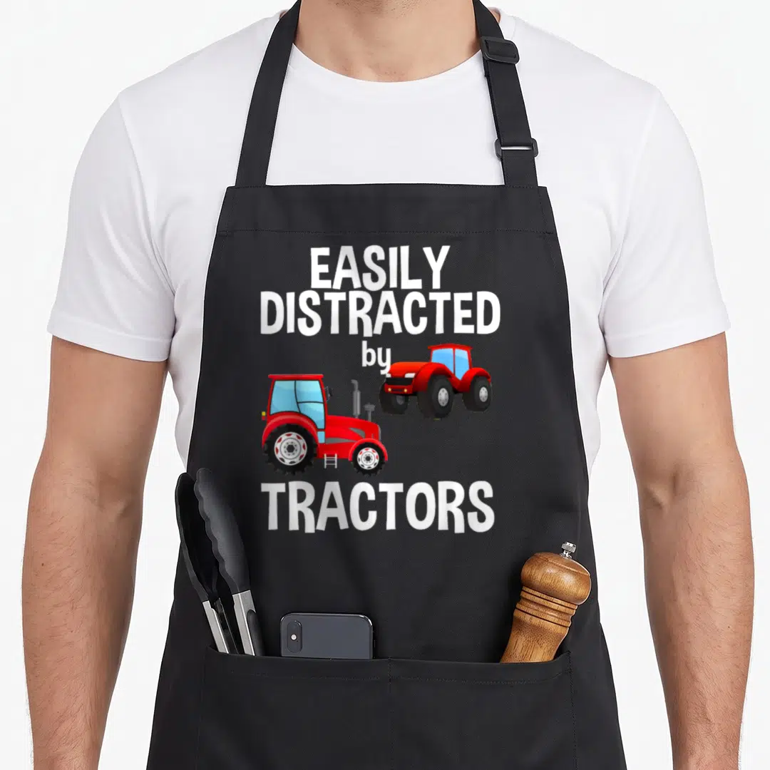 Easily farmer apron - Two red cartoon tractors below stacked white text EASILY DISTRACTED by TRACTORS — shown on a black cotton canvas apron against a white background