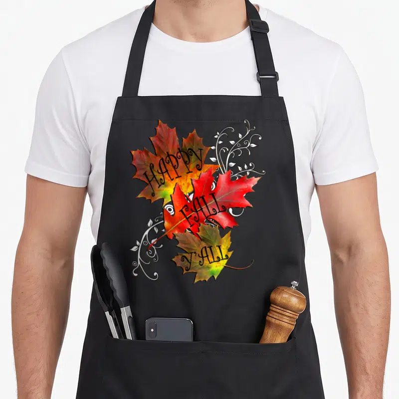 Pattern fall aprons - Three stacked maple leaves in red orange yellow green with HAPPY FALL Y ALL text and white vines — shown on a black cotton canvas apron against a white background