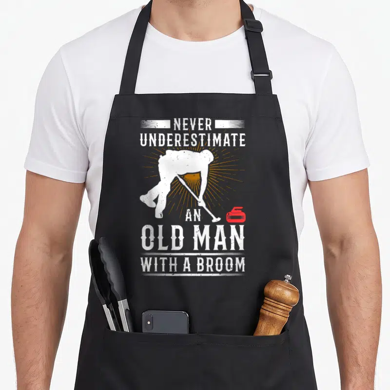 Underestimate curling apron - Distressed white text surrounding a curler silhouette, orange sunburst, and red stone — shown on a black cotton canvas apron against a white background