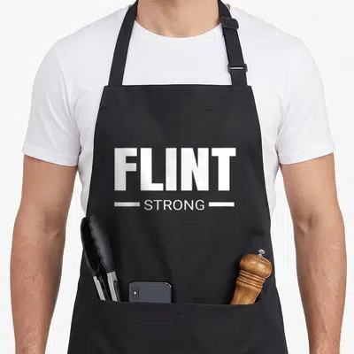 Strong flint apron - White text FLINT STRONG stacked with horizontal lines on black fabric — shown on a black cotton canvas apron against a white background