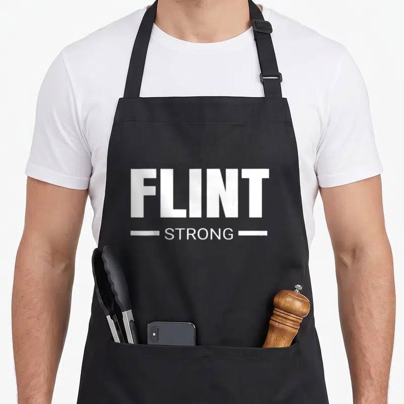 Strong flint apron - White text FLINT STRONG stacked with horizontal lines on black fabric — shown on a black cotton canvas apron against a white background