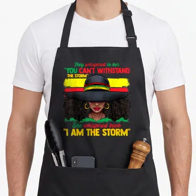 History storm african apron - Cartoon woman with wide hat and 'I AM THE STORM' text on tri-color striped background — shown on a black cotton canvas apron against a white background