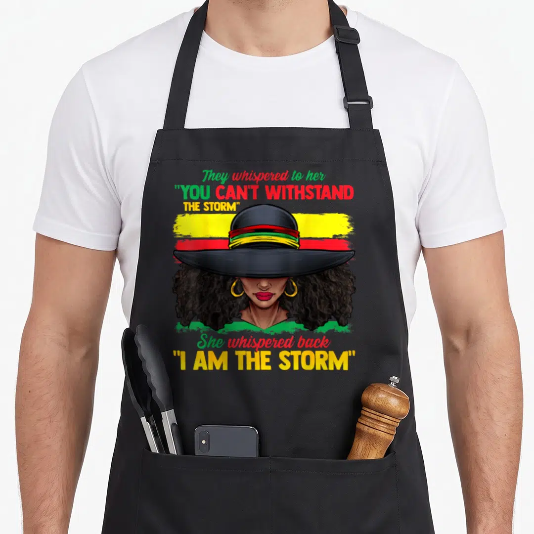 History storm african apron - Cartoon woman with wide hat and 'I AM THE STORM' text on tri-color striped background — shown on a black cotton canvas apron against a white background