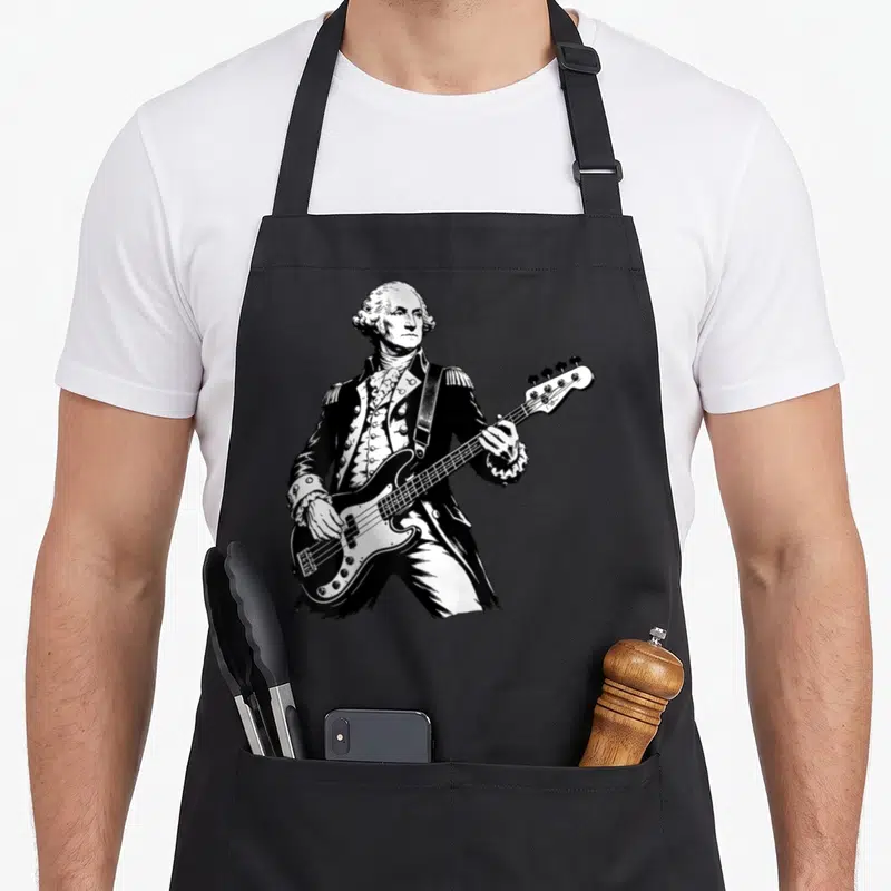 Guitar 4th of july apron - Black and white line art of a man in colonial uniform playing a bass guitar — shown on a black cotton canvas apron against a white background