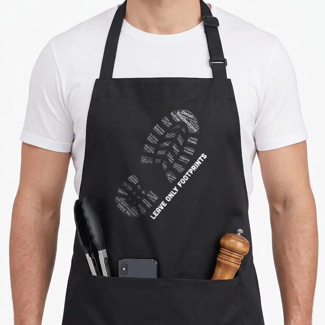 National police lover apron - White text forming a boot shape listing US National Park names and Leave Only Footprints — shown on a black cotton canvas apron against a white background