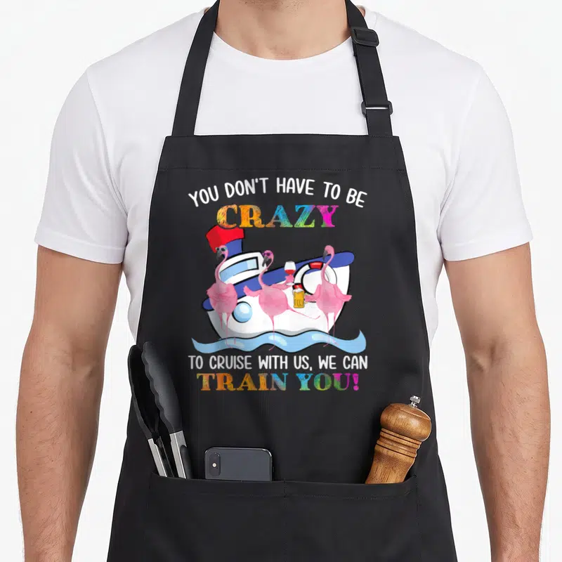 Cruise flamingo apron - Three pink flamingos holding drinks on a cartoon cruise ship with rainbow text — shown on a black cotton canvas apron against a white background