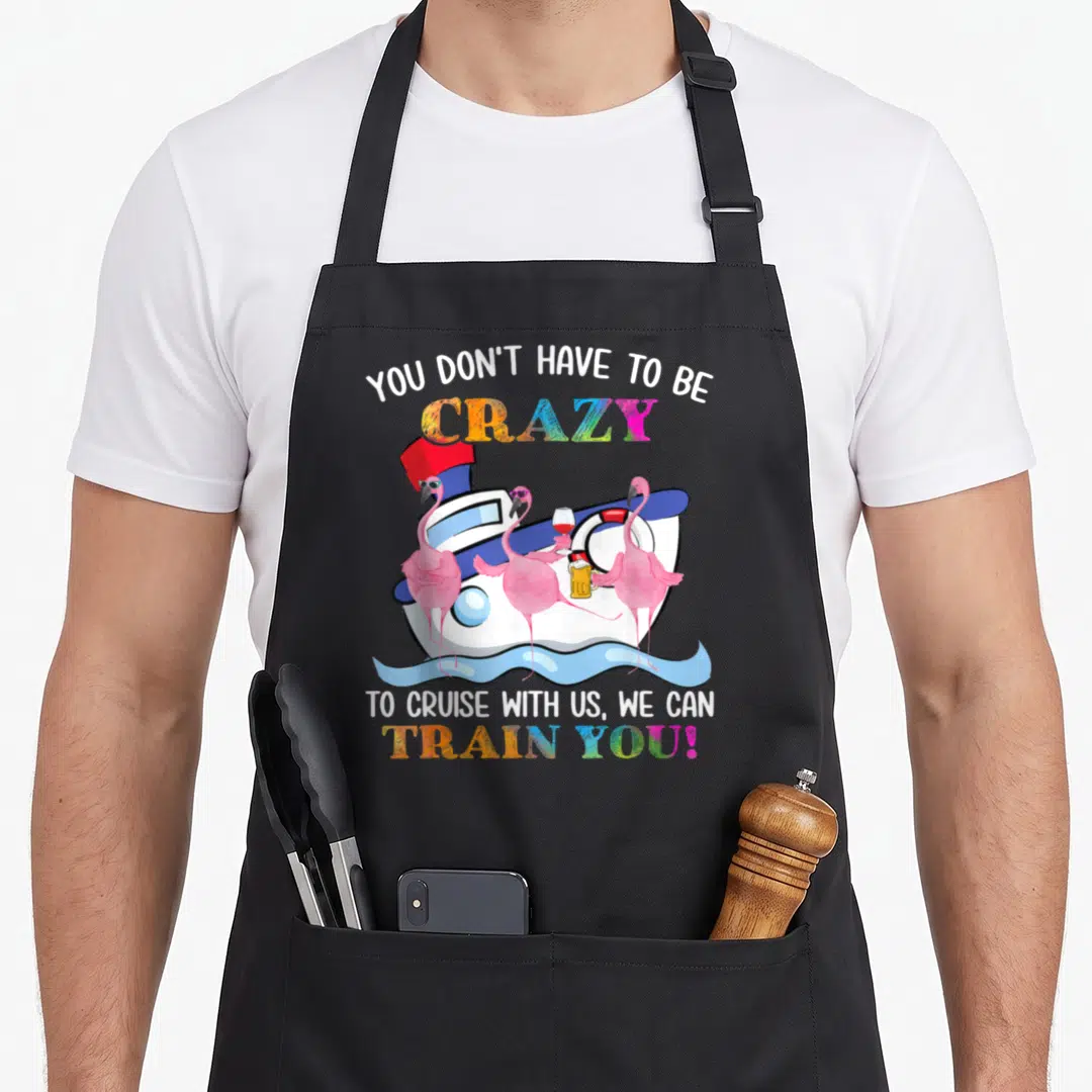 Cruise flamingo apron - Three pink flamingos holding drinks on a cartoon cruise ship with rainbow text — shown on a black cotton canvas apron against a white background