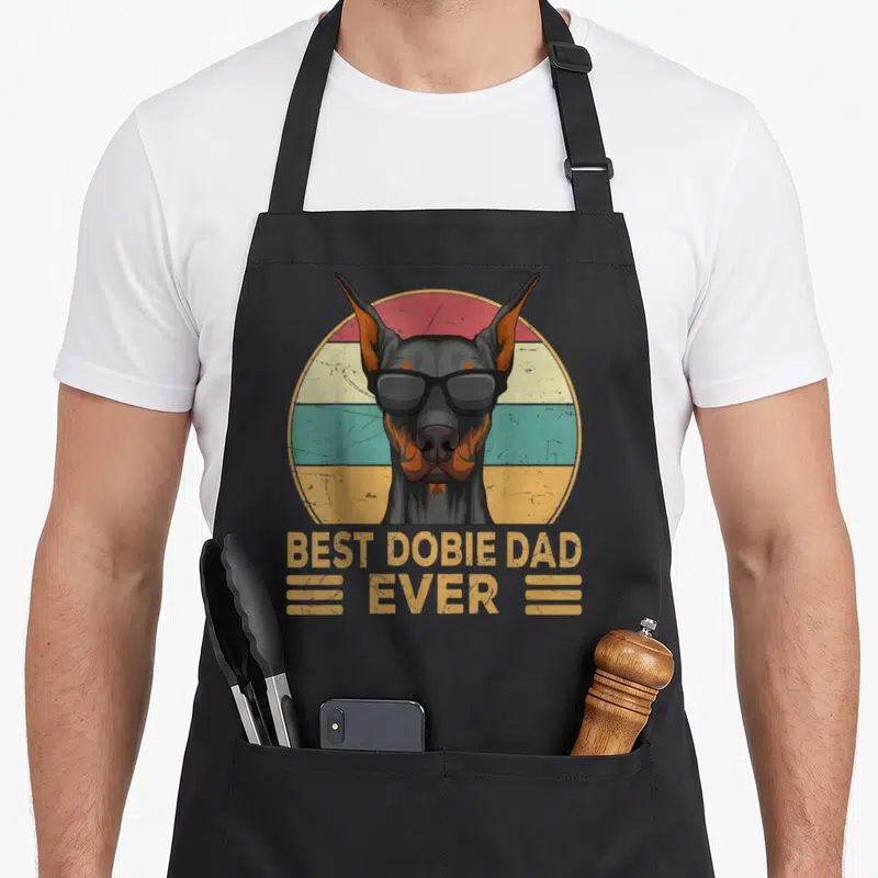 Dobermann dog dad apron - Retro Doberman in sunglasses with BEST DOBIE DAD EVER text on striped circle — shown on a black cotton canvas apron against a white background