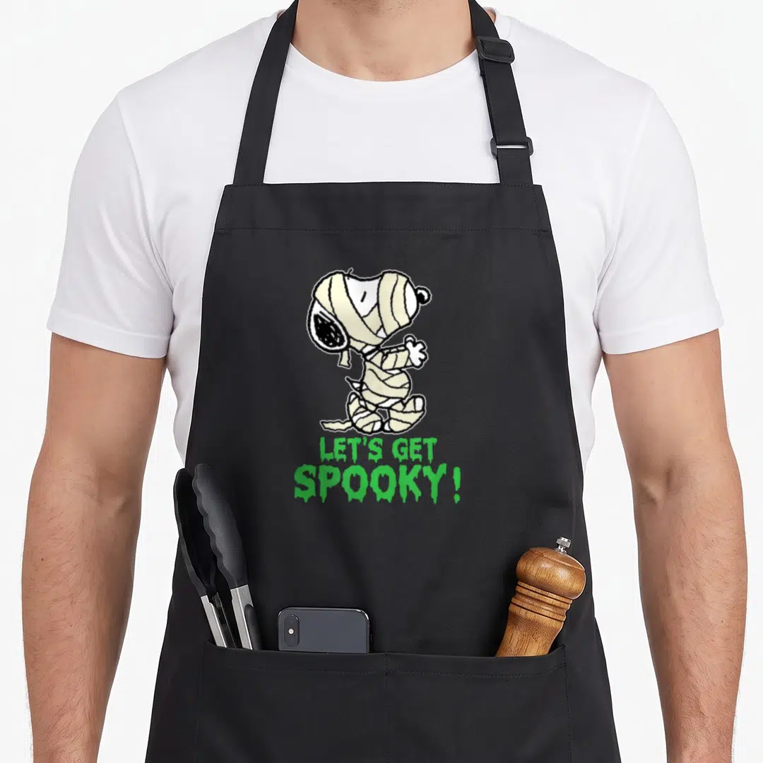 Funny spooky apron - Snoopy wrapped in beige mummy bandages with green LET'S GET SPOOKY text below — shown on a black cotton canvas apron against a white background