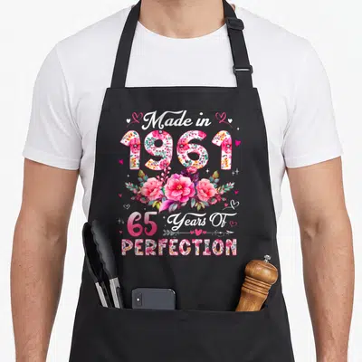 Funny 65th vintage floral apron - Floral filled 1961 and 65 text with pink flowers and hearts celebrating a 65th — shown on a black cotton canvas apron against a white background