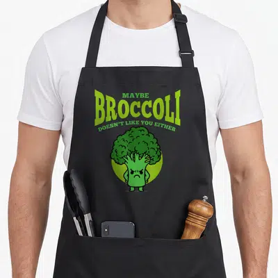 Maybe vegan apron - Angry cartoon broccoli character with green and lime text reading Maybe Broccoli Doesn't Like You — shown on a black cotton canvas apron against a white background