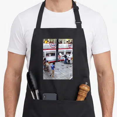 Riverfront montgomery apron - Photo of people fighting near a white tram with red and blue bunting on a wooden dock — shown on a black cotton canvas apron against a white background