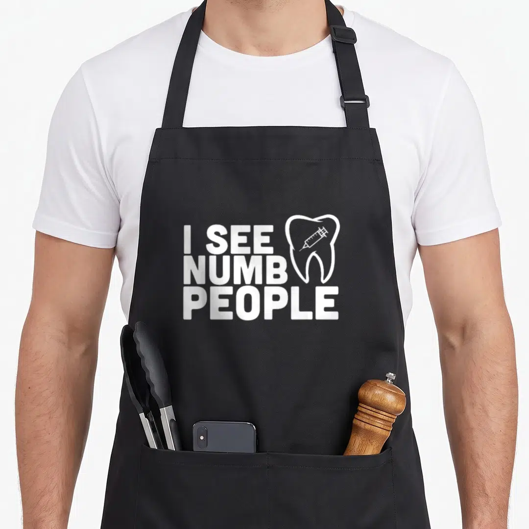 See dentist apron - White text I See Numb People with tooth and syringe icon — shown on a black cotton canvas apron against a white background