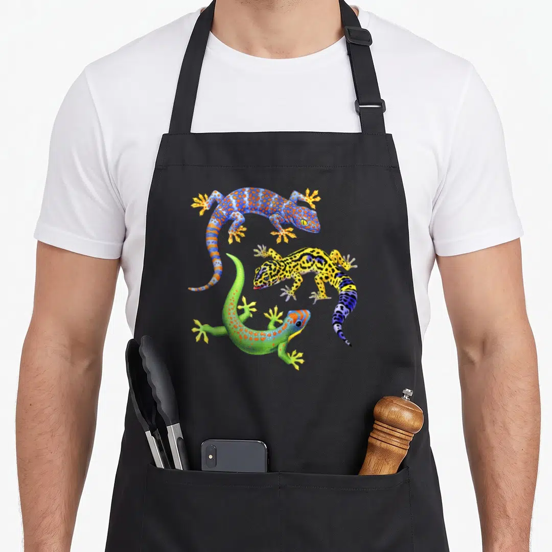 Wildlife gecko apron - Three realistic geckos in blue, yellow, green, and orange spotted patterns — shown on a black cotton canvas apron against a white background