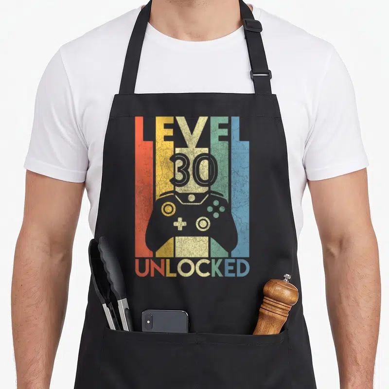 Unlocked birthday apron - Retro game controller with LEVEL 30 UNLOCKED text over red orange yellow green blue grey — shown on a black cotton canvas apron against a white background