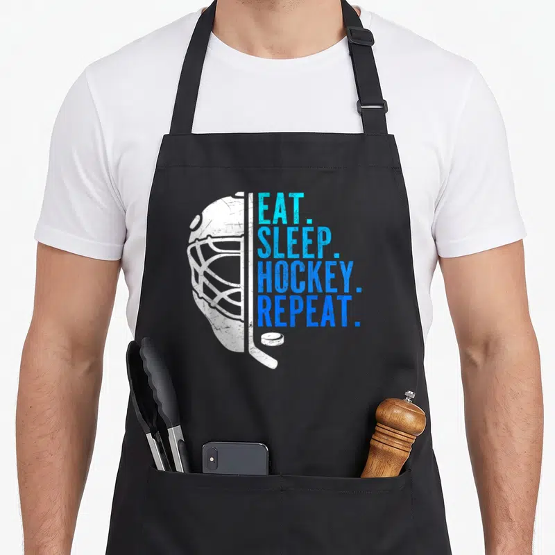 Eat hockey ice apron - White distressed hockey helmet with blue stacked text EAT SLEEP HOCKEY REPEAT on black bib — shown on a black cotton canvas apron against a white background