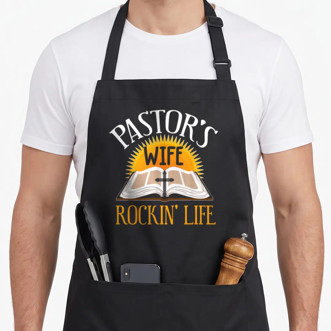 Pastor family aprons - Cartoon sun and open bible with cross and PASTOR'S WIFE ROCKIN' LIFE text — shown on a black cotton canvas apron against a white background