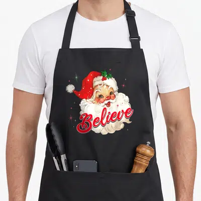 Believe christmas santa claus apron - Watercolor Santa head with red Believe script and holly leaves — shown on a black cotton canvas apron against a white background