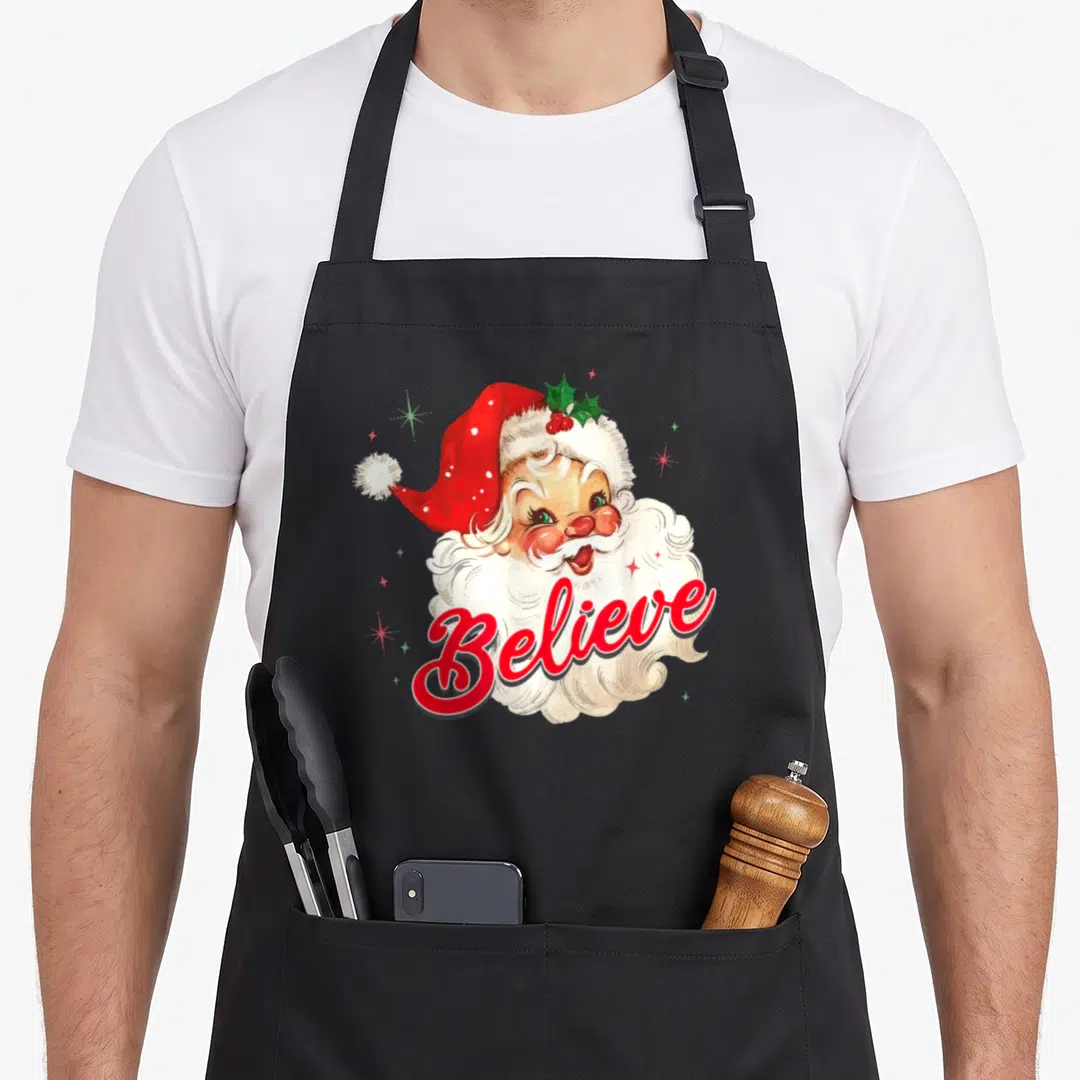 Believe christmas santa claus apron - Watercolor Santa head with red Believe script and holly leaves — shown on a black cotton canvas apron against a white background