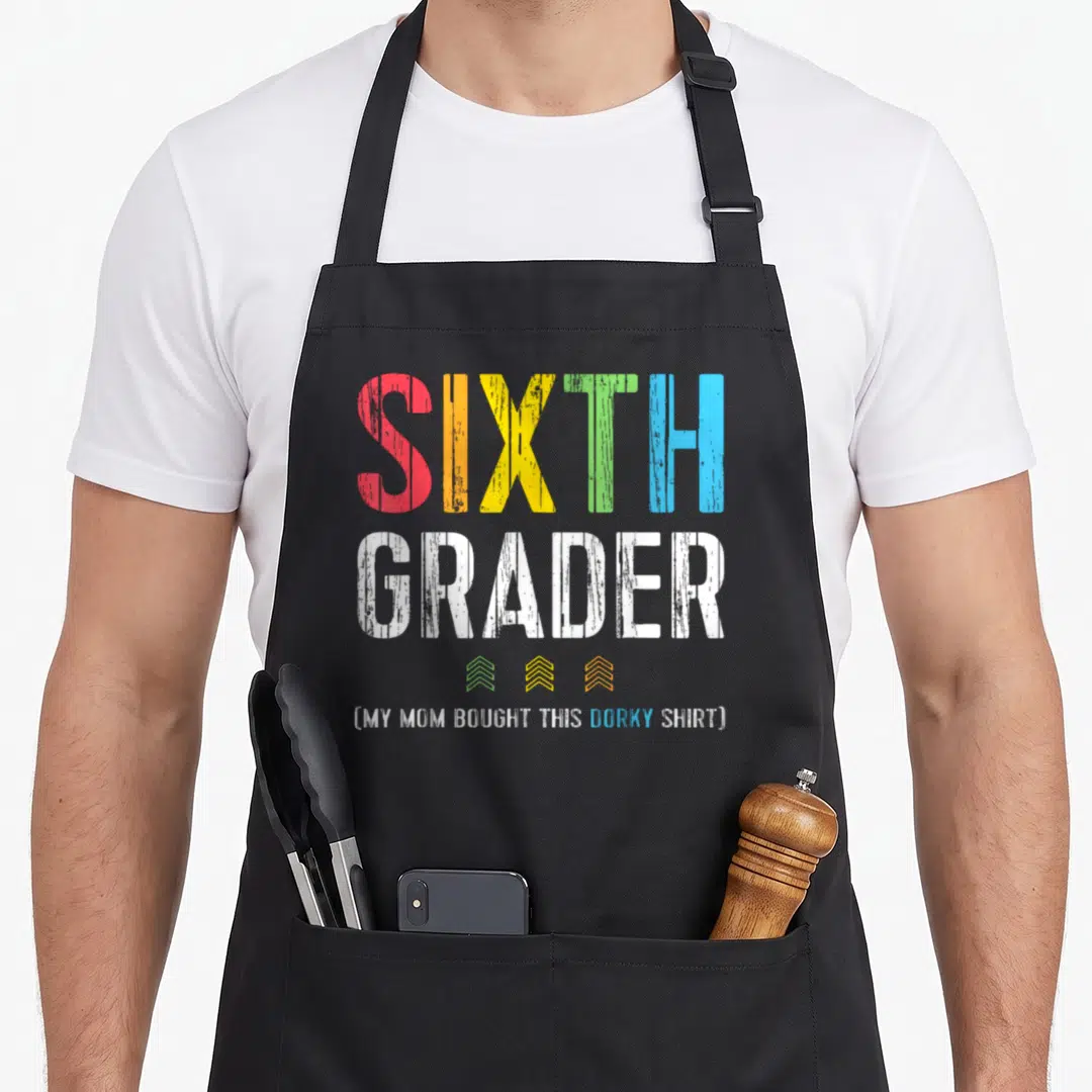 6th grade apron - Stacked colorful text reading SIXTH GRADER with three small chevrons and parenthetical joke below — shown on a black cotton canvas apron against a white background