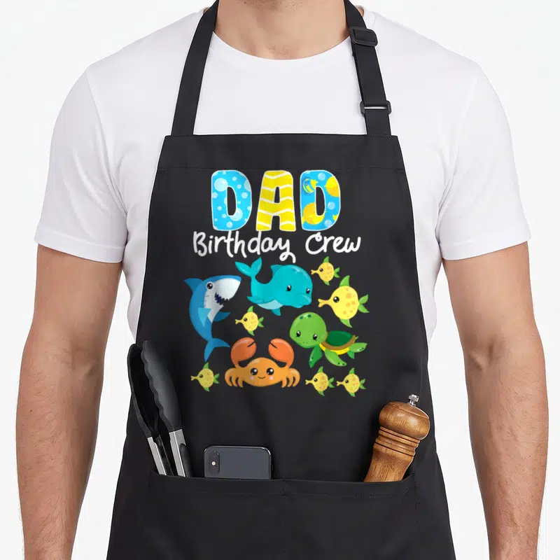Birthday fish apron - DAD Birthday Crew text with cartoon shark, dolphin, turtle, crab, and yellow fish in bright colors — shown on a black cotton canvas apron against a white background