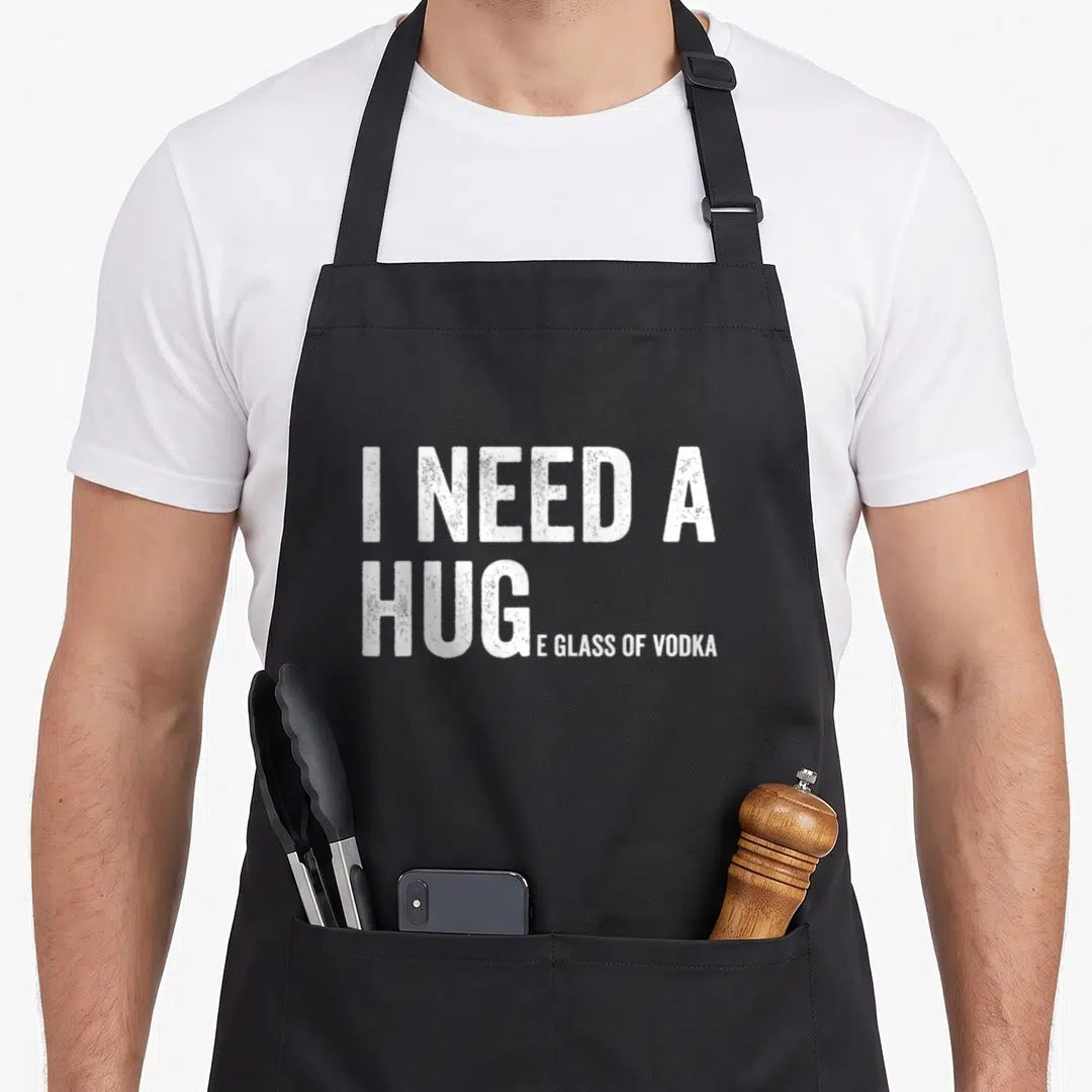 Huge glass humor apron - White distressed text I NEED A HUG...E GLASS OF VODKA on grey background — shown on a black cotton canvas apron against a white background