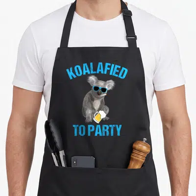 Koalafied animal apron - Grey koala with blue sunglasses holding yellow beer mug and cigarette — shown on a black cotton canvas apron against a white background