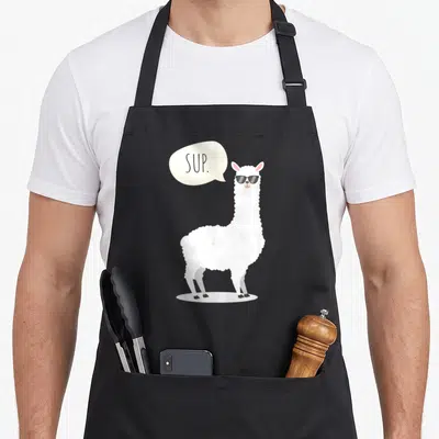 Sup alpaca apron - White cartoon llama in black sunglasses with beige speech bubble saying SUP — shown on a black cotton canvas apron against a white background