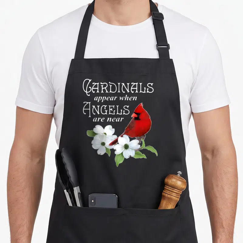 Appear cardinal apron - Red cardinal perched on white dogwood flowers below stacked white text reading Cardinals appear — shown on a black cotton canvas apron against a white background
