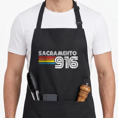 Proud sacramento apron - Retro Sacramento 916 design with red orange yellow blue stripes and EST 1850 text — shown on a black cotton canvas apron against a white background