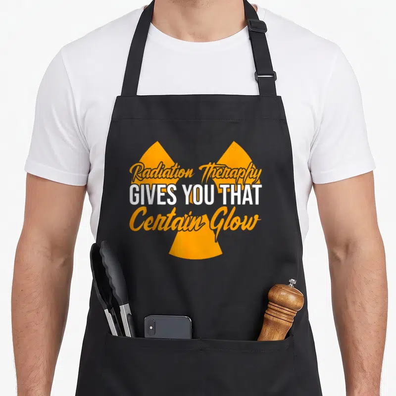 Give radiation aprons - Orange radiation symbol with white and orange text reading Radiation Therapy Gives You That — shown on a black cotton canvas apron against a white background