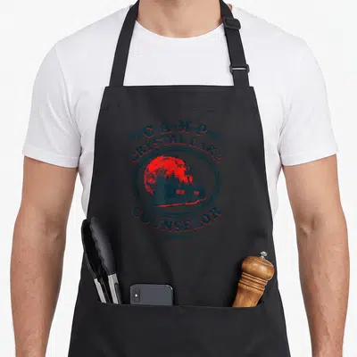 Camp counselor apron - Distressed blue text Camp Crystal Lake Counselor surrounding red moon and cabin illustration — shown on a black cotton canvas apron against a white background