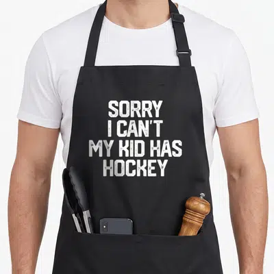 Sorry hockey dad apron - White distressed text reading SORRY I CAN'T MY KID HAS HOCKEY stacked vertically — shown on a black cotton canvas apron against a white background