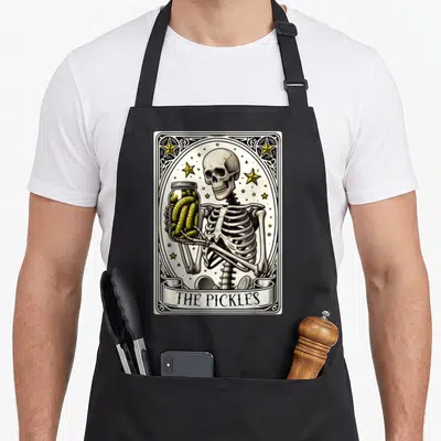 Tarot canning apron - Skeleton holding pickle jar inside ornate frame with banner text THE PICKLES — shown on a black cotton canvas apron against a white background