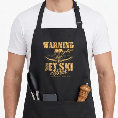 Jet ski apron - Tan distressed skull on jet ski with WARNING Certified JET SKI Addict text — shown on a black cotton canvas apron against a white background
