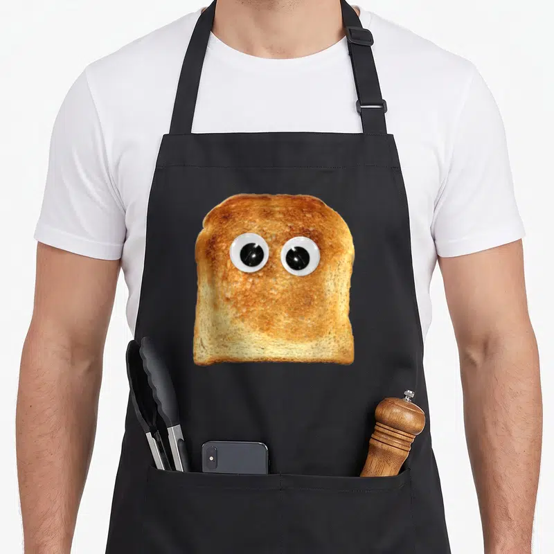 Googly toast apron - Photo of brown toast with two large white and black googly eyes centered on bread — shown on a black cotton canvas apron against a white background