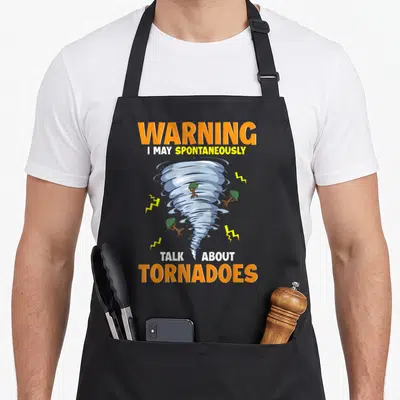 May talk apron - Cartoon tornado with scattered trees lightning bolts and stacked warning text in orange blue yellow — shown on a black cotton canvas apron against a white background
